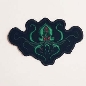Puede incluir: Parche bordado en negro y verde con un pulpo estilizado con ojos rojos. El pulpo está rodeado de un fondo negro.