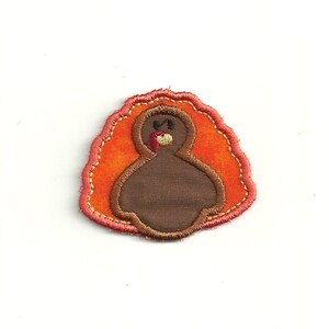 May include: A brown felt turkey applique with a red wattle and orange felt feathers. The turkey is outlined in gold thread.