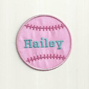 May include: A pink baseball patch with embroidered details. The patch features the name "Hailey" in teal thread across the center. Pink stitching outlines the baseball shape and mimics the seams. The patch is circular and sits on a white background.