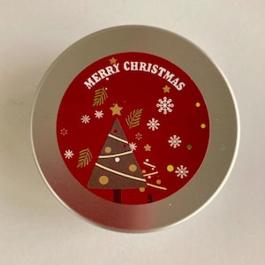 May include: A circular, silver tin with a red label displaying "MERRY CHRISTMAS". The label features a brown Christmas tree, snowflakes, stars, and decorative leaves. The tin is likely for a festive, holiday-themed product.