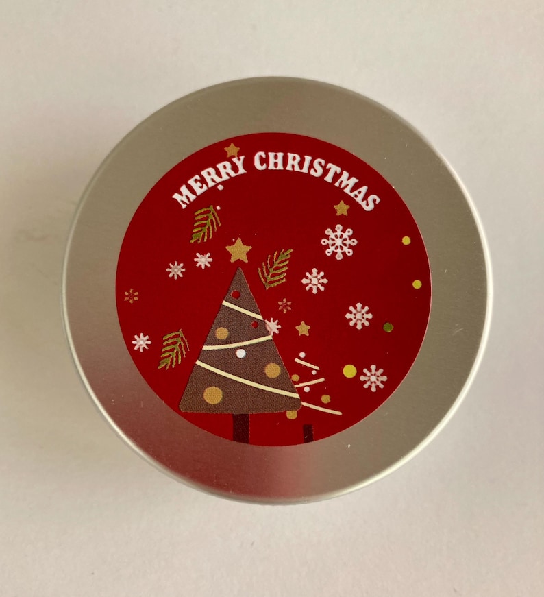 May include: A circular, silver tin with a red label displaying "MERRY CHRISTMAS". The label features a brown Christmas tree, snowflakes, stars, and decorative leaves. The tin is likely for a festive, holiday-themed product.