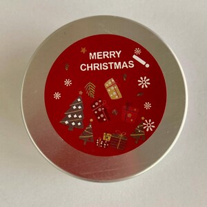 May include: A circular, silver container with a red label displaying the words "MERRY CHRISTMAS". The label is adorned with white snowflakes, Christmas trees, and gift illustrations. The container is likely for a festive product.
