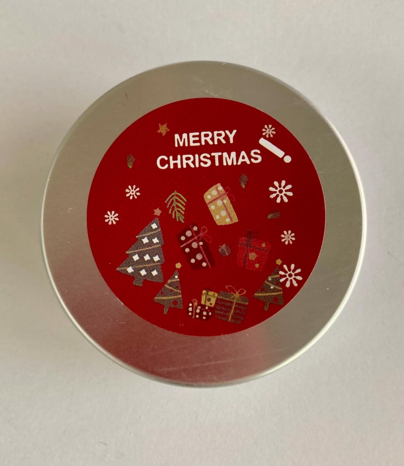 May include: A circular, silver container with a red label displaying the words "MERRY CHRISTMAS". The label is adorned with white snowflakes, Christmas trees, and gift illustrations. The container is likely for a festive product.