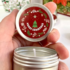 May include: A small, silver candle tin with a red lid. The lid has a Christmas tree design and the words "Merry Christmas." White snowflakes, leaves, and a bow decorate the lid. The candle inside is white.