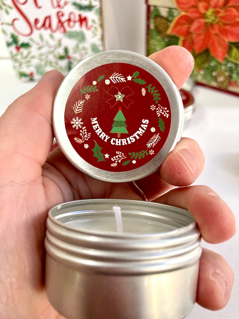 May include: A small, silver candle tin with a red lid. The lid has a Christmas tree design and the words "Merry Christmas." White snowflakes, leaves, and a bow decorate the lid. The candle inside is white.