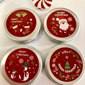 May include: Four small, round, silver tins with red lids featuring Christmas designs. The lids show images of Santa Claus, snowmen, bells, and a Christmas tree, with the words "Merry Christmas". A red and white candy is also visible.