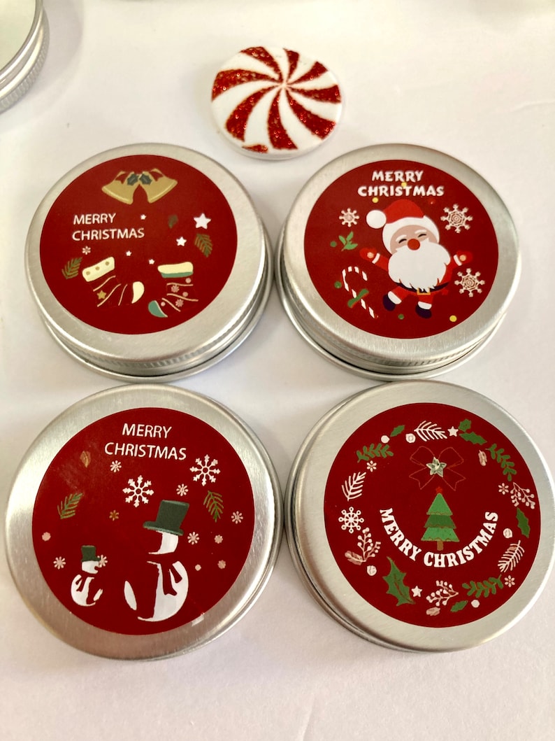 May include: Four small, round, silver tins with red lids featuring Christmas designs. The lids show images of Santa Claus, snowmen, bells, and a Christmas tree, with the words "Merry Christmas". A red and white candy is also visible.