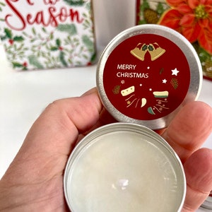 May include: A small, round, silver tin with a red lid featuring the words "MERRY CHRISTMAS" and festive illustrations. The tin is open, revealing a white candle. A holiday-themed gift box and card are in the background.