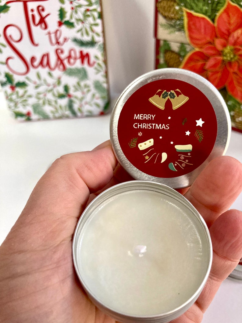 May include: A small, round, silver tin with a red lid featuring the words "MERRY CHRISTMAS" and festive illustrations. The tin is open, revealing a white candle. A holiday-themed gift box and card are in the background.