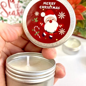 May include: A small, silver candle tin with a white candle. The lid has a red label with a Santa Claus illustration and the words "MERRY CHRISTMAS". A Christmas card is visible in the background.