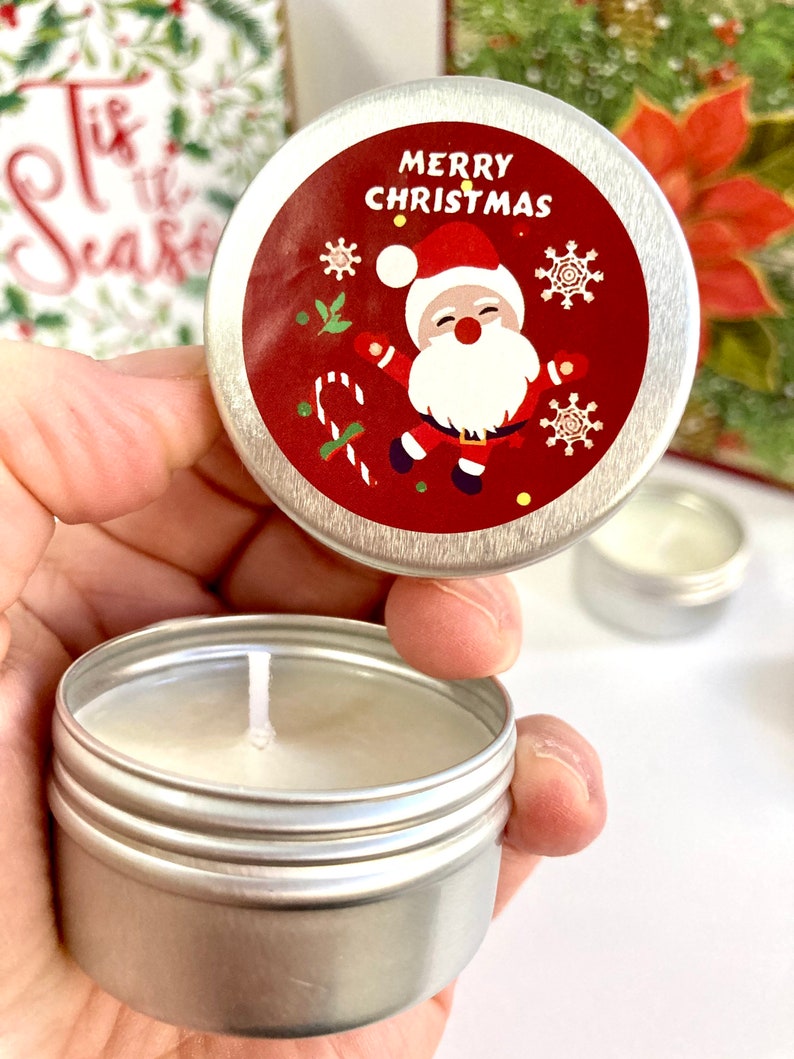 May include: A small, silver candle tin with a white candle. The lid has a red label with a Santa Claus illustration and the words "MERRY CHRISTMAS". A Christmas card is visible in the background.