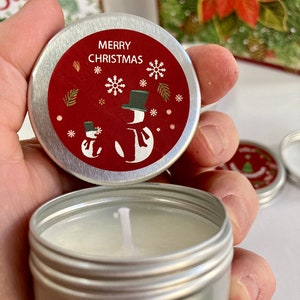 May include: A small, silver candle tin with a red lid. The lid has the text "MERRY CHRISTMAS", snowflakes, and snowman illustrations. The candle inside is white, and the wick is visible. The theme is Christmas.