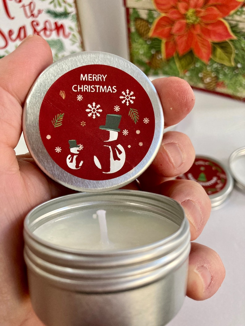 May include: A small, silver candle tin with a red lid. The lid has the text "MERRY CHRISTMAS", snowflakes, and snowman illustrations. The candle inside is white, and the wick is visible. The theme is Christmas.