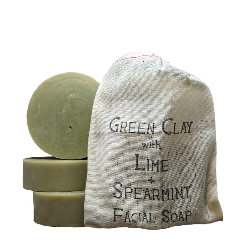 May include: Four green clay facial soap bars stacked on top of each other. The soap bars are in a white muslin bag with the text "GREEN CLAY with LIME + SPEARMINT FACIAL SOAP" printed on it.