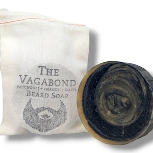 Op de afbeelding: Een ronde, zwart-bruine baardzeep met een swirl-patroon. De zeep is verpakt in een witte trekkoordtas met de tekst "THE VAGABOND PATCHOULI + ORANGE + CLOVE BEARD SOAP" en een tekening van een baard.