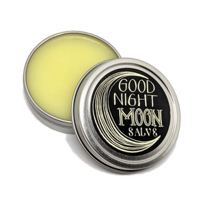 May include: A silver tin with a black label that reads "Good Night Moon Salve". The label features a stylized crescent moon with a white outline. The tin is open and a yellow salve is visible inside.