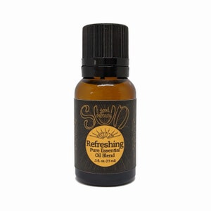 Pode incluir: Um pequeno frasco de vidro âmbar com uma tampa preta e um rótulo preto. O rótulo tem um gráfico de sol e o texto "good morning SUN Refreshing Pure Essential Oil Blend 5 fl. oz. (15 ml)"