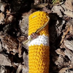 May include: A yellow and white crocheted pouch with a brown bead and a green cord. The pouch is lying on a bed of brown wood chips.