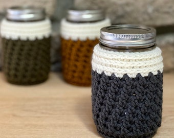 Peut inclure: Trois bocaux en verre avec des couvercles en crochet. Les bocaux sont en verre transparent avec des couvercles argentés. Les couvercles sont crochetés dans différentes couleurs : vert foncé, orange et gris. Le couvercle gris a une bande blanche autour du haut.