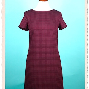 Könnte beinhalten: Ein kurzes, burgunderfarbenes Kleid mit kurzen Ärmeln und einem strukturierten Muster. Das Kleid befindet sich auf einer Puppe vor einem türkisfarbenen Hintergrund.