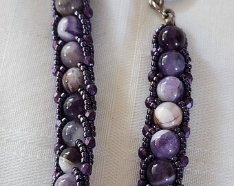 May include: A purple and white beaded bracelet with a silver clasp. The beads are arranged in a braided pattern.