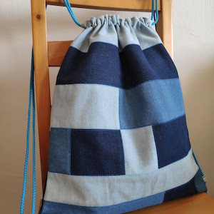 Op de afbeelding: Een blauwe en lichtblauwe denim trekkoordtas met een patchwork ontwerp. De tas hangt aan een houten stoel.