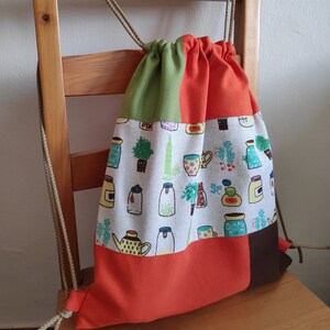 May include: A drawstring bag made of patchwork fabric. The bag is orange, green, brown, and white. The white fabric has a pattern of jars, cups, and plants. The bag is hanging on a wooden chair.