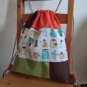 May include: A drawstring bag made of patchwork fabric. The bag has a green, brown, white, and orange design. The white fabric features a pattern of jars, teapots, and plants.