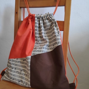 Può includere: Borsa a cordoncino arancione, marrone e bianca con un motivo a note musicali. La borsa è realizzata in tessuto e ha una chiusura a cordoncino.