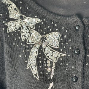 May include: Black cardigan with silver sequin bow detailing. The bows are adorned with clear sequins and beads. The sweater features black buttons and a round neckline.