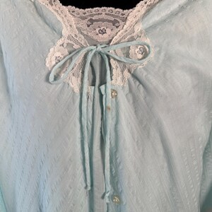 May include: A light blue vintage robe with a textured pattern. The robe features a white lace collar and trim, with a matching tie at the neckline. The front has a button closure. The sleeves are short, and the overall style is delicate and feminine.
