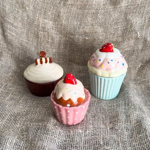 May include: Three ceramic cupcake containers with colourful frosting and sprinkles. The cupcakes are in different colours: brown, pink and blue.