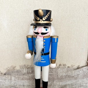 May include: A wooden nutcracker figurine with a blue uniform, white pants, black boots, and a black hat with gold trim. The nutcracker has a white beard and mustache, a red nose, and blue eyes.