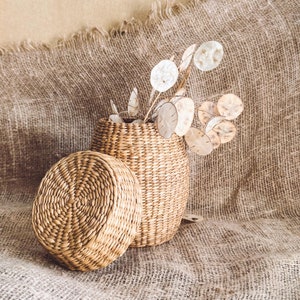 May include: A woven wicker basket with a lid, filled with dried white flowers, sitting on a brown burlap surface.