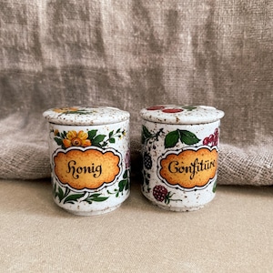 Peut inclure: Deux petits pots en céramique blanche avec couvercles, décorés de motifs floraux et de baies. L'un porte l'inscription "Honig" sur une étiquette orange, l'autre "Confiture". Les pots ont une finition mouchetée et sont posés sur un fond neutre.