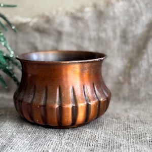 Può includere: Un vaso di rame con un design scanalato. Il vaso è appoggiato su una superficie di juta.