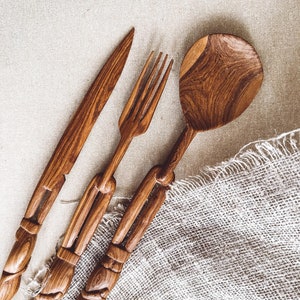 Könnte beinhalten: Ein Set aus drei Holzbesteckteilen, bestehend aus Messer, Gabel und Löffel, mit aufwendigen Schnitzereien. Das Besteck ist aus dunkelbraunem Holz gefertigt und hat ein rustikales, handgemachtes Aussehen.