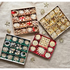 May include: Four square boxes containing assorted straw ornaments in various shapes like stars, wreaths, and cones. The boxes feature dividers and include natural straw-coloured items, some accented with small red or blue ribbons and bows.
