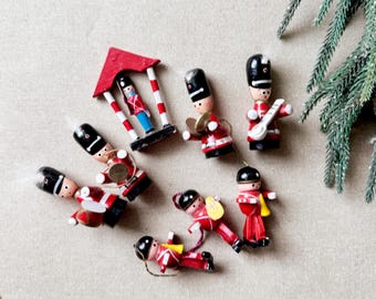 May include: A collection of miniature wooden toy soldiers in red and black uniforms, some playing instruments, others standing or lying down. A red and white striped structure with a soldier inside is also present. A sprig of evergreen adds a festive touch.