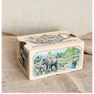 Könnte beinhalten: Eine rechteckige Holzkiste mit Rich Brew Tea, Fine Ceylon High Grown BOP Tea, Nettogewicht 200 Gramm. Die Oberseite zeigt eine Teekannen-Illustration, während die Vorderseite eine farbenfrohe Szene mit einem Elefanten und Menschen zeigt.
