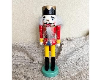 May include: A wooden nutcracker figurine with a red and yellow uniform, a black hat with a gold crown, a white beard, and blue eyes. The nutcracker is standing on a green base.