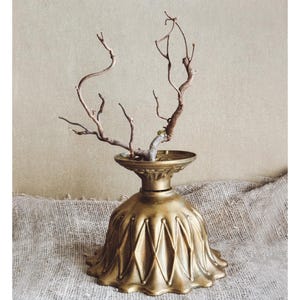 Pode incluir: Um vaso decorativo de latão vintage com uma base canelada em forma de sino e um pescoço fino, segurando galhos secos. O vaso tem um acabamento dourado antigo e quente e fica em uma superfície texturizada de tons neutros.