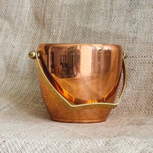 May include: A copper ice bucket with a brass handle. The bucket is round and has a shiny, reflective surface.