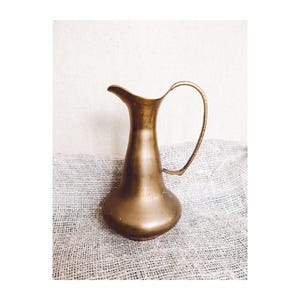 May include: A vintage brass jug with a curved handle and a spout. The jug has a bulbous base and a slender neck. It is displayed on a textured, grey fabric background.