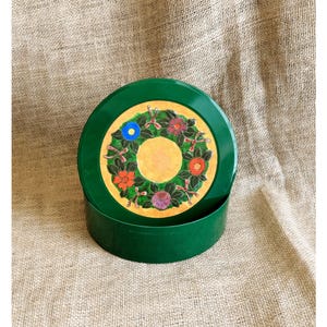 Pode incluir: Uma lata redonda de metal verde-escuro com uma tampa apresentando um círculo amarelo central rodeado por uma coroa decorativa de folhas verdes, pequenos laços cor-de-rosa e flores coloridas em tons de azul, vermelho, laranja e roxo sobre ouro.