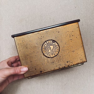 Puede incluir: Una caja rectangular de metal dorado con un patrón texturizado en forma de rejilla. La caja tiene una parte superior negra y un emblema circular en la parte delantera. El emblema presenta un diseño ornamentado con texto. La caja es sostenida por una mano.