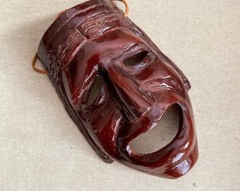 Pode incluir: Uma máscara teatral castanho-avermelhada brilhante com uma boca larga e aberta e orifícios para os olhos. A máscara tem um design detalhado e esculpido nas bordas e um cordão de couro para pendurar. A máscara é provavelmente usada para apresentações teatrais.