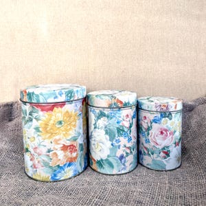 May include: Three cylindrical metal storage canisters of varying sizes, each featuring a colourful floral pattern with yellow, orange, pink, and blue flowers against a light background. Each canister includes a matching removable lid.