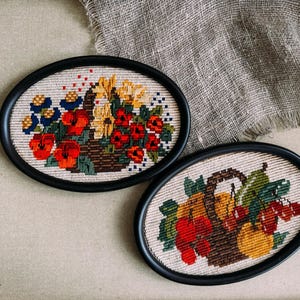 May include: Two framed embroidery pieces with a floral and fruit design. The top piece features a basket of red flowers and yellow flowers. The bottom piece features a basket of red and yellow fruit.
