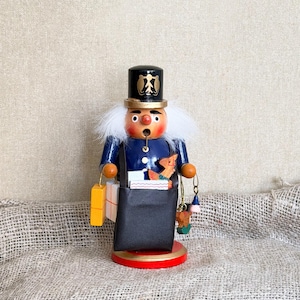 Puede incluir: Una figura de cascanueces de madera con sombrero de copa negro con detalles dorados, chaqueta azul y pelo blanco. El personaje lleva un bolso de hombro negro lleno de cajitas de regalo y un animal de madera sobre una base circular roja.